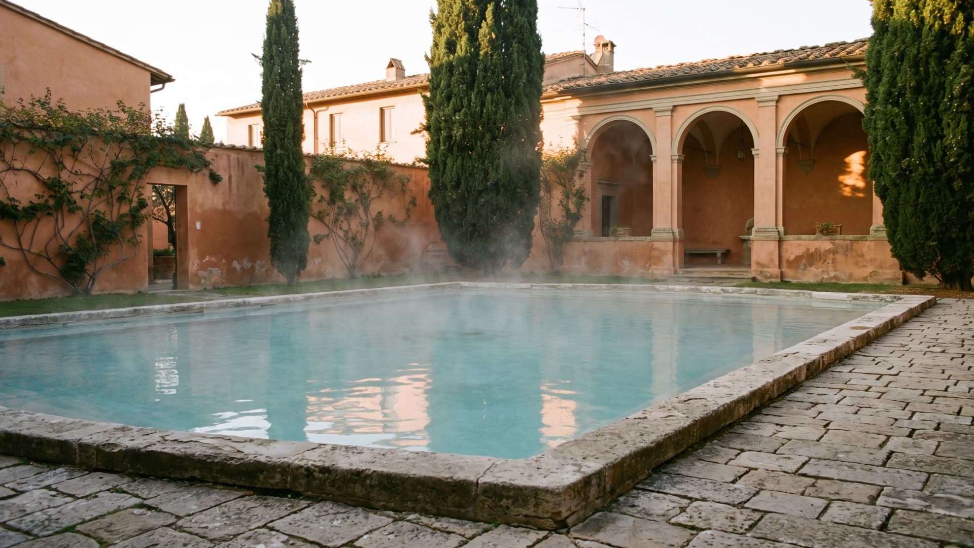 A stone thermal pool in a quiet Italian courtyard with terracotta walls and cypress trees, steam drifting over the water