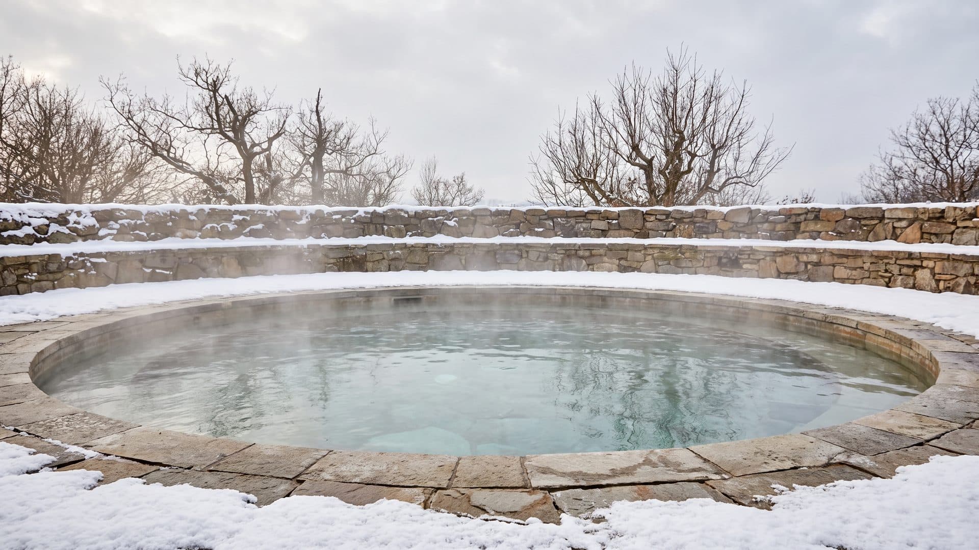 Una piscina termal al aire libre humeando en el aire frío, con nieve sobre el brocal de piedra y el muro de detrás, y árboles desnudos bajo un cielo gris.