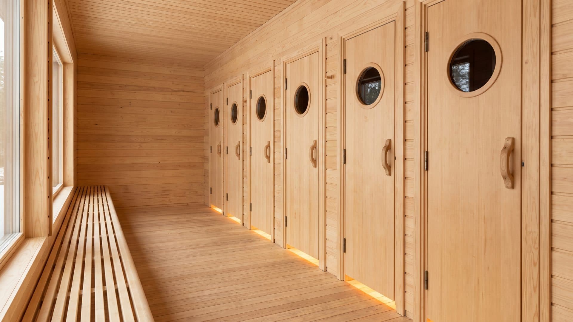 Una hilera de sencillas puertas de sauna de madera a lo largo de un pasillo silencioso, cada una con un pequeño ojo de buey, y un banco de listones enfrente.