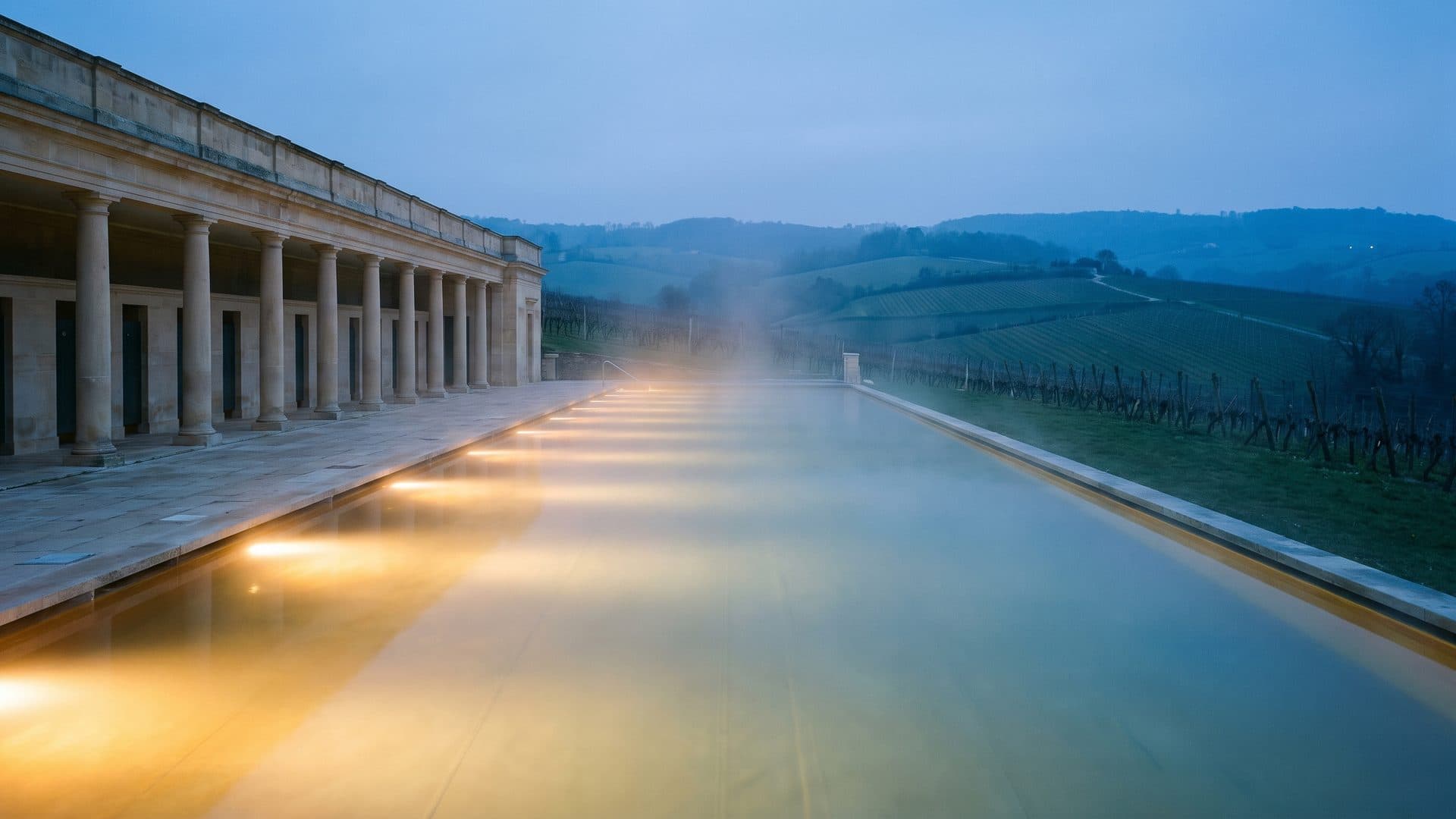 A long outdoor thermal pool at dusk, lit from below, with a colonnaded bath building along one side and vineyard hills fading behind