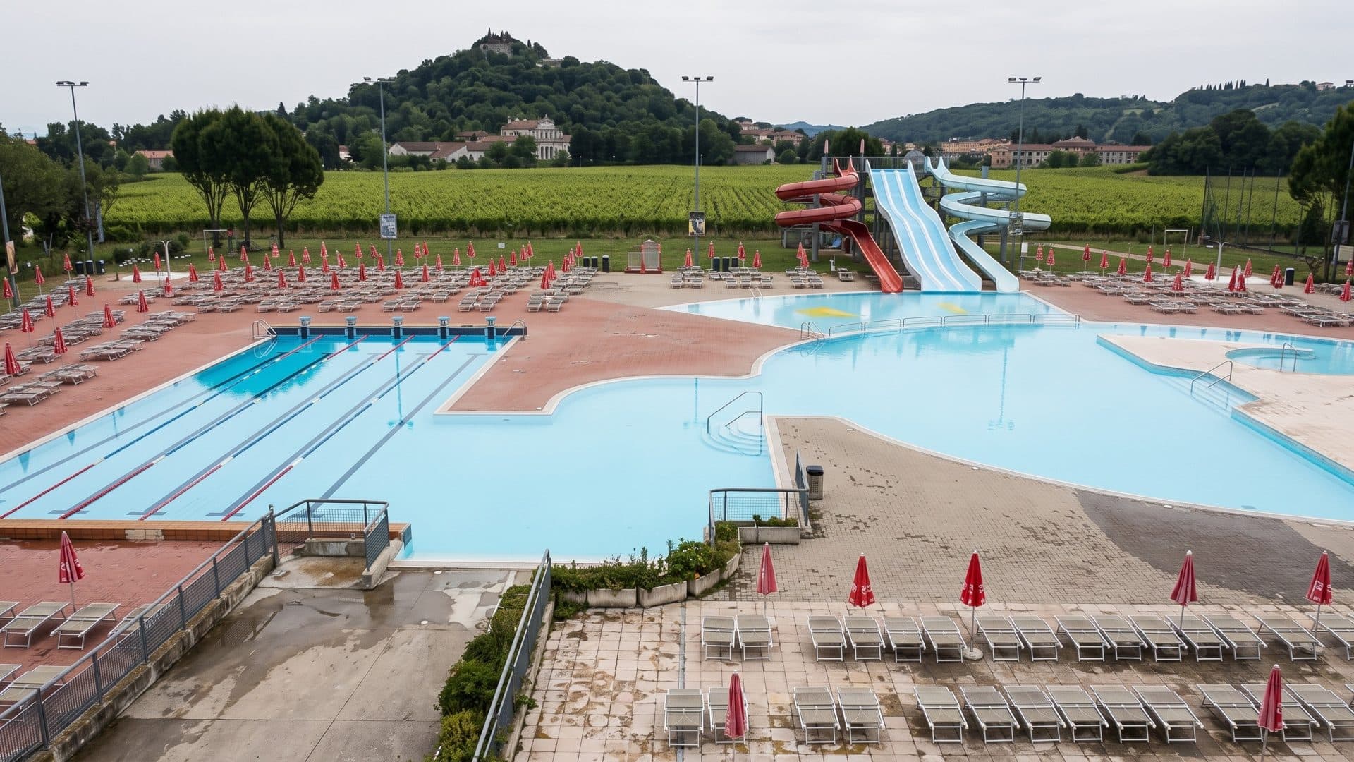 The outdoor pools of Acquapark Altavilla Vicentina with slides and red parasols, hills behind