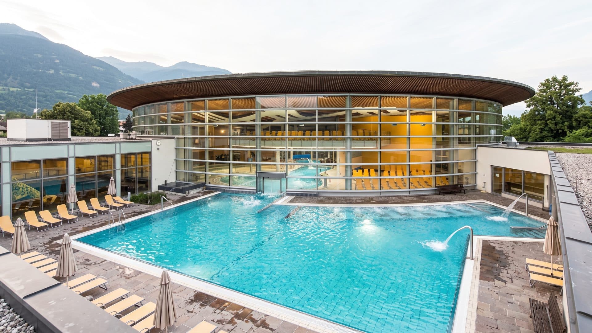 Outdoor pool in front of the curved timber and glass hall of the baths, mountains behind.