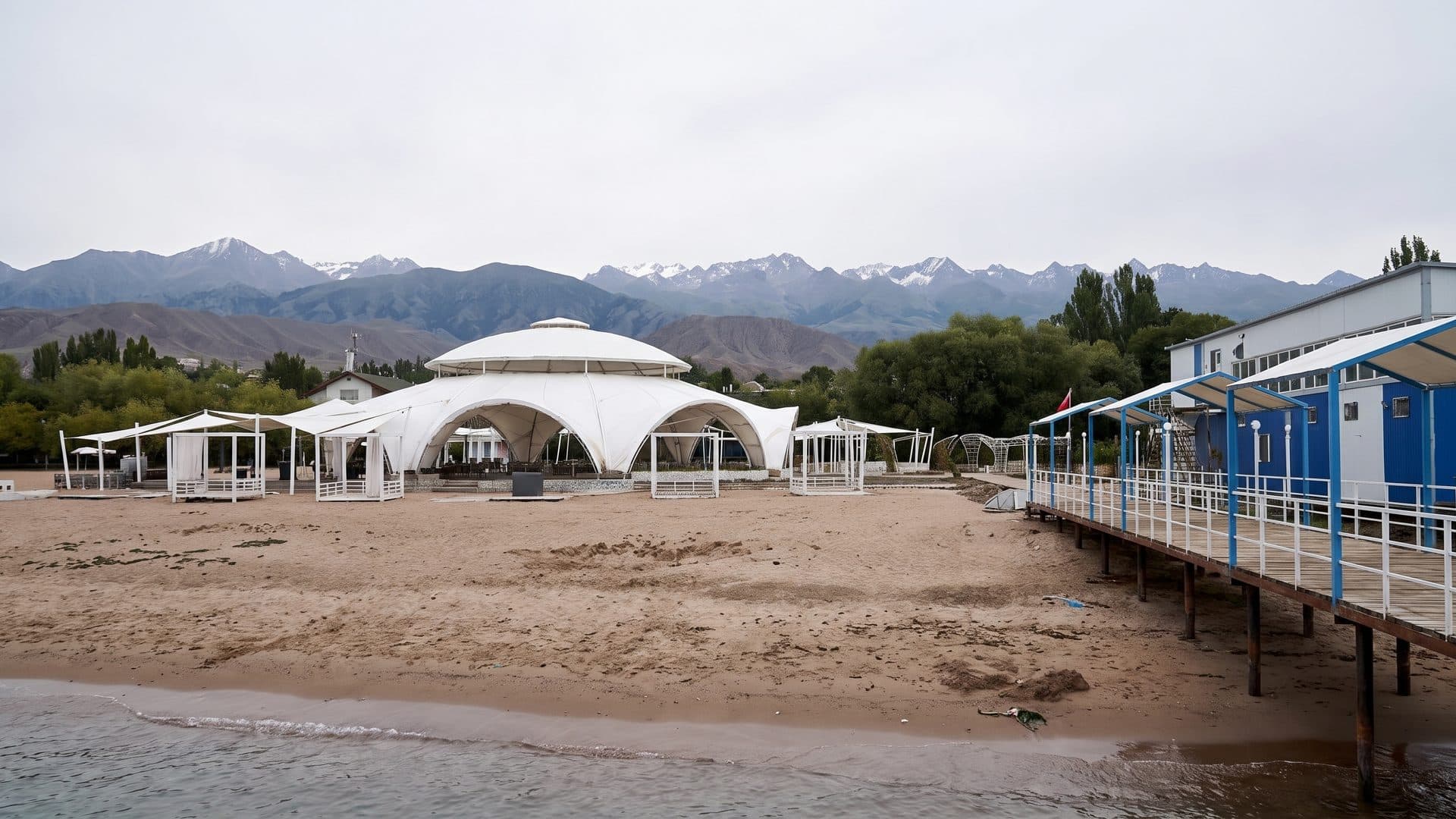 The beach at Ak-Bermet on Lake Issyk-Kul, with a white marquee pavilion, a timber pier and blue changing cabins under the Tien Shan mountains