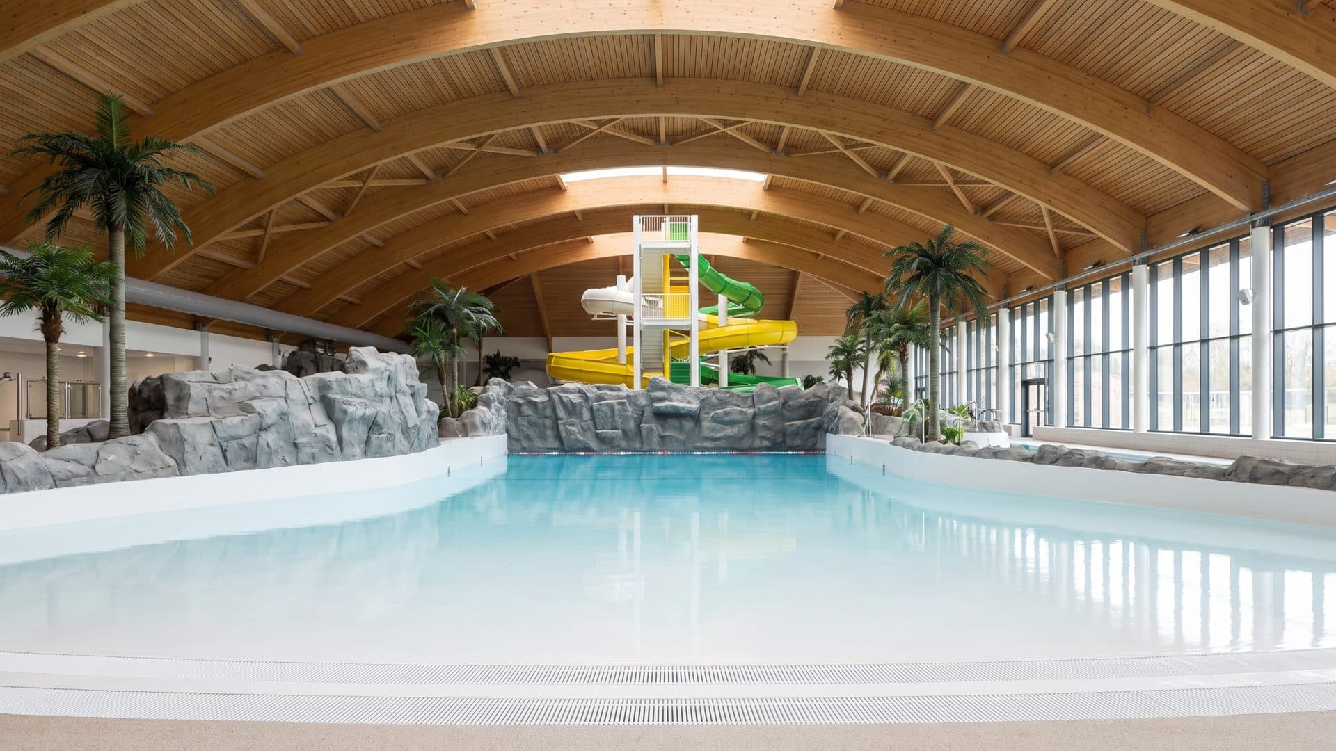 The pool of the Akvamir water park below its coloured slide tower under the curved timber roof