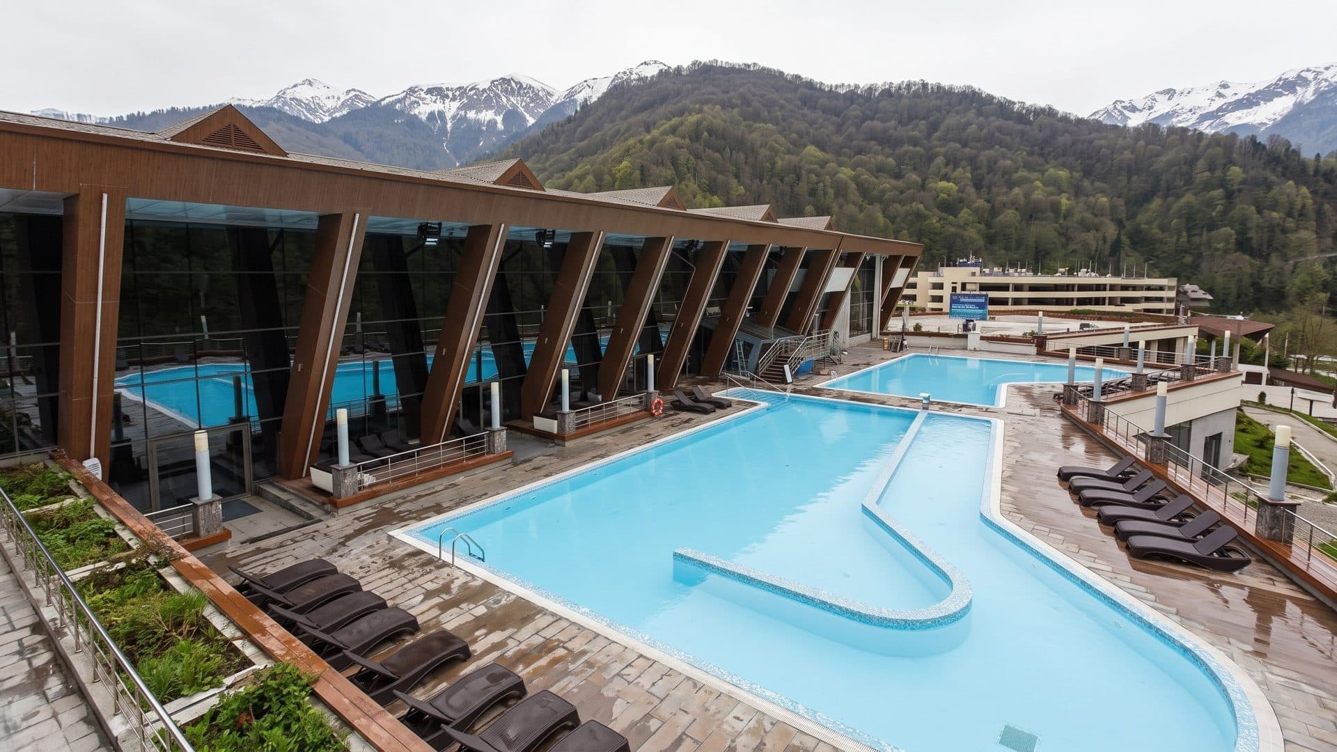 The outdoor pool of the Galaktika water park beside a timber-framed glass gallery, forested mountains behind