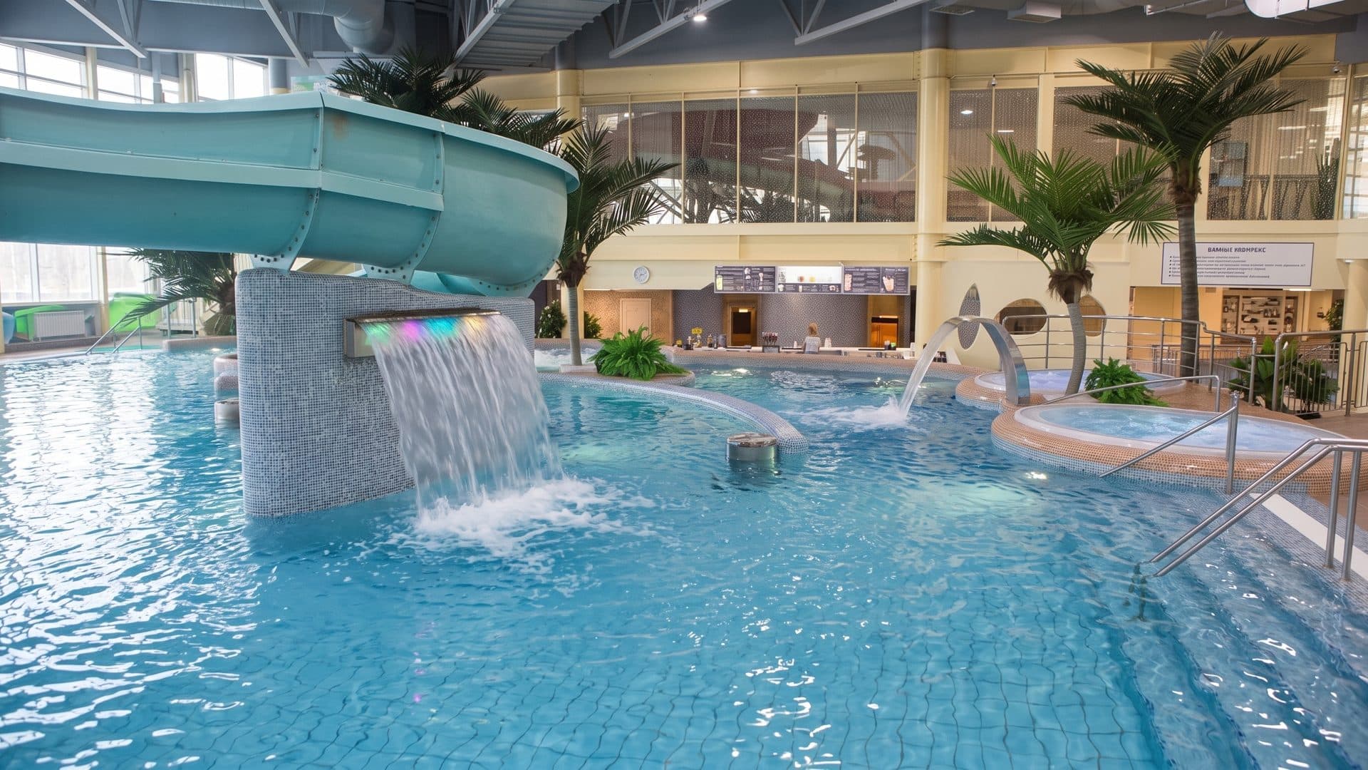The indoor leisure pool of Akvapolis in Pskov with a waterfall under a slide, a whirlpool and palms