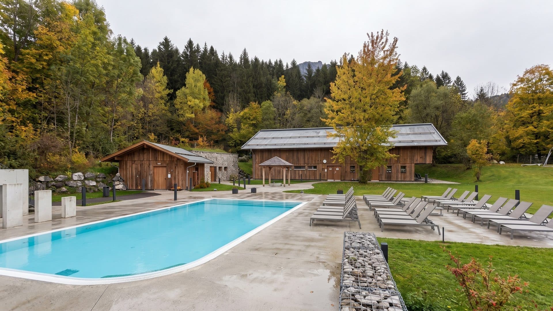 Outdoor thermal pool on a paved terrace, with timber sauna huts and autumn larches on the wooded slope behind