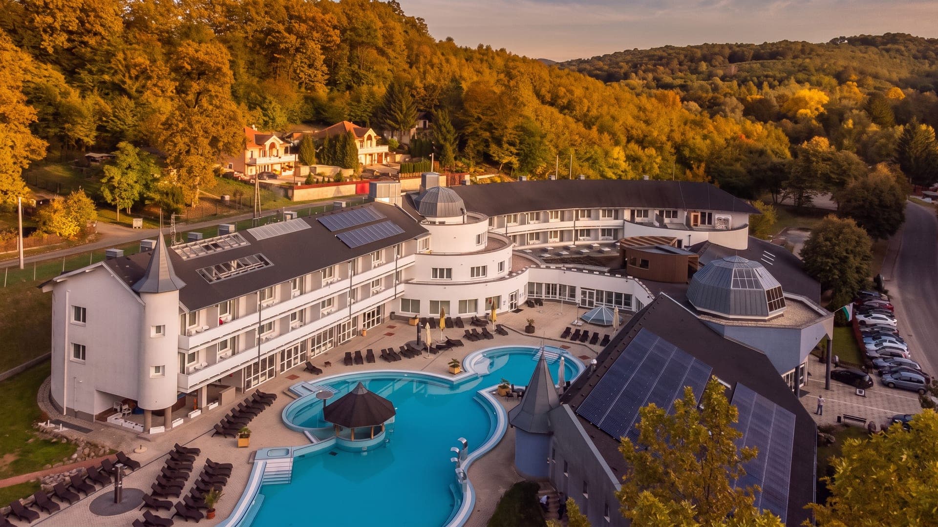 Aerial view of the white spa hotel and its curved outdoor pools, surrounded by wooded autumn hills