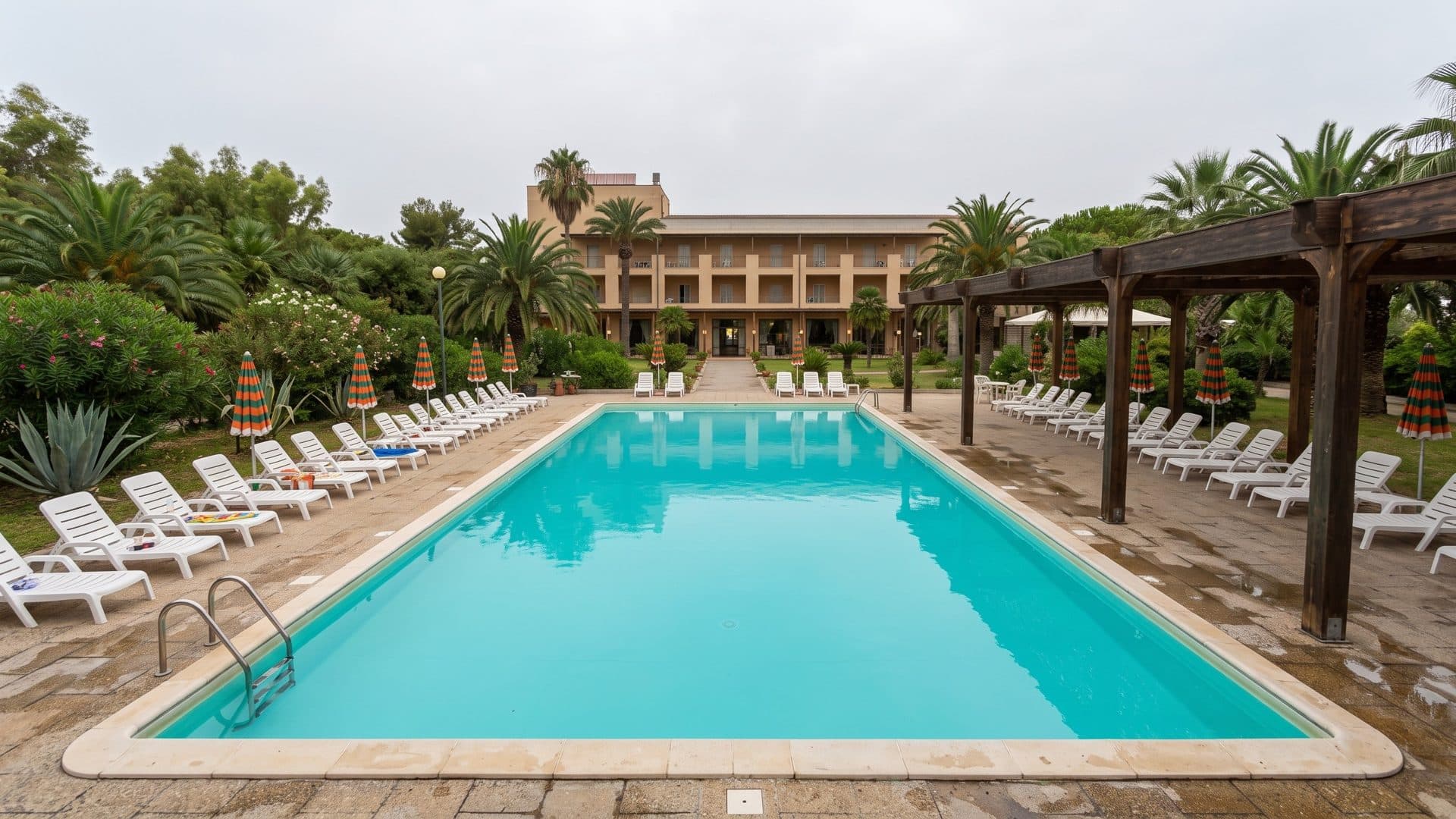 The rectangular outdoor pool of the Antiche Terme di Sardara lined with white loungers under a pergola and palm trees, the hotel behind