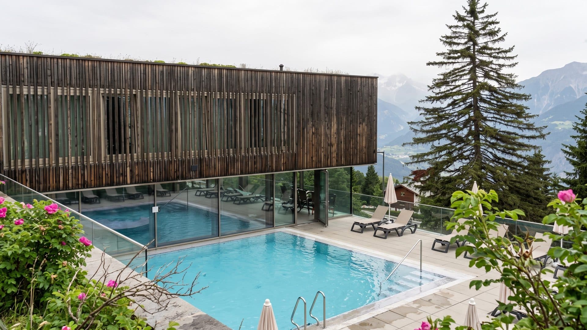 The timber-clad Anzere spa building above its glass-walled pool hall and an outdoor pool, fir trees and Alpine peaks around
