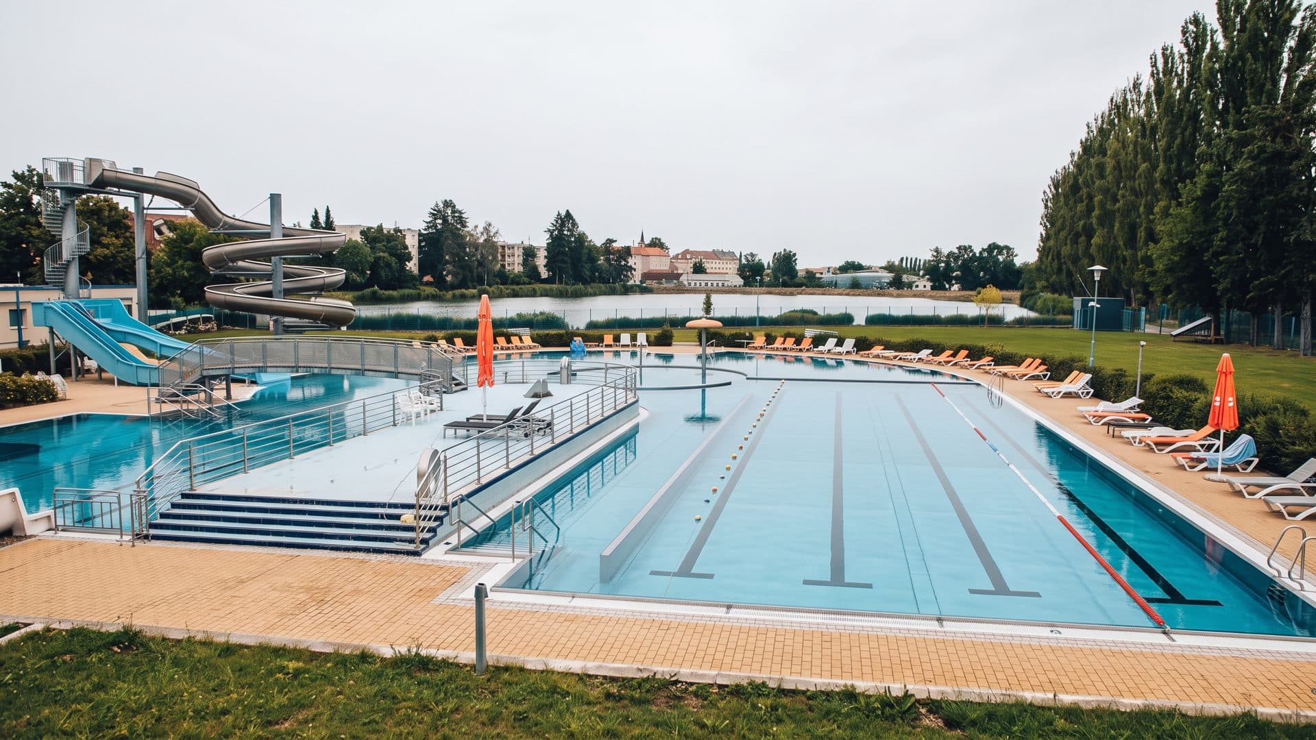 The outdoor lane pool of Aqua Centrum Jicin with slides beside it, poplars and the pond behind