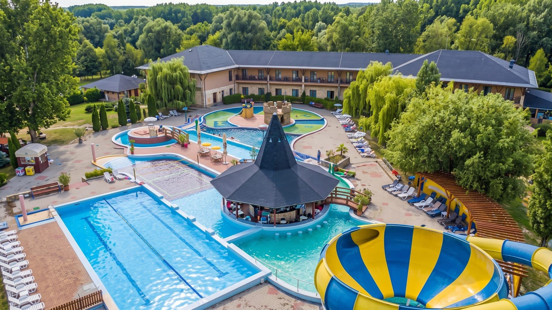 Outdoor pools of the thermal park from the air: swimming pool, curved leisure basins, a conical thatched pavilion and a blue and yellow slide.