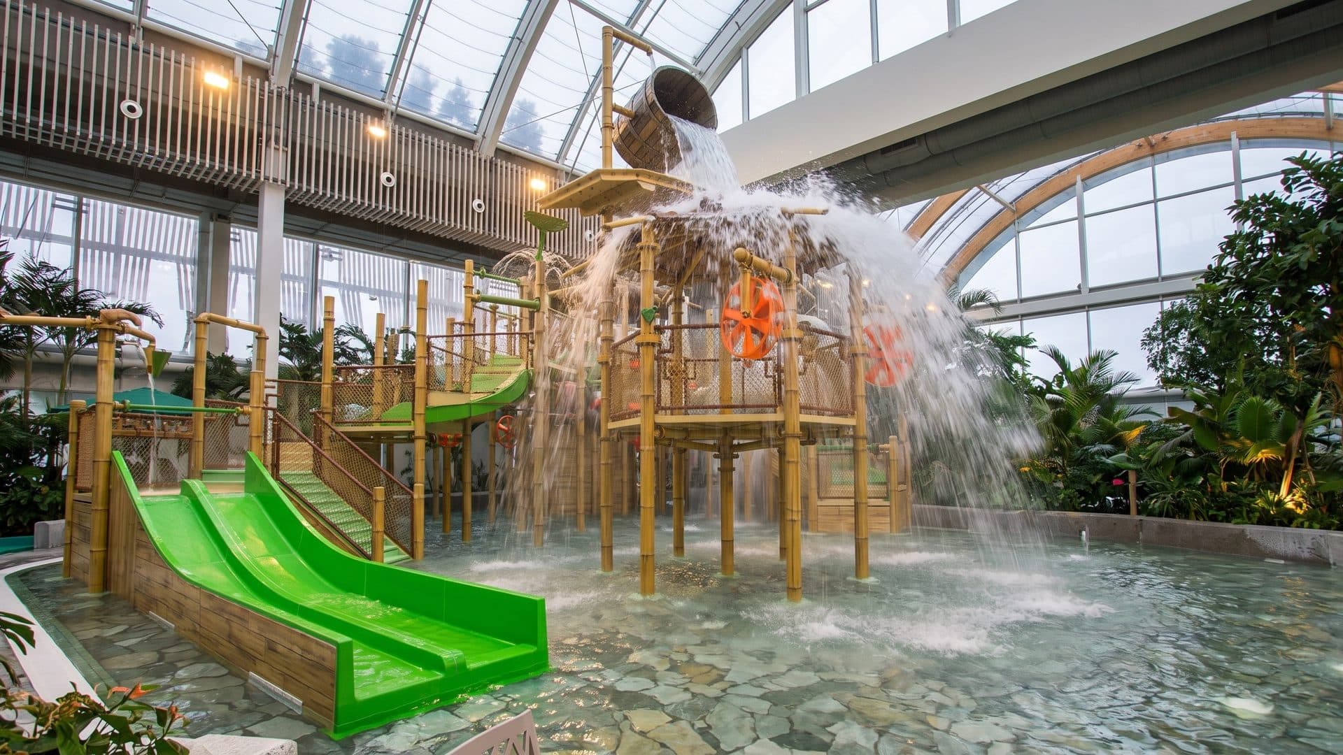 Indoor water playground under a glass roof, a timber climbing tower with green slides and a tipping bucket pouring water