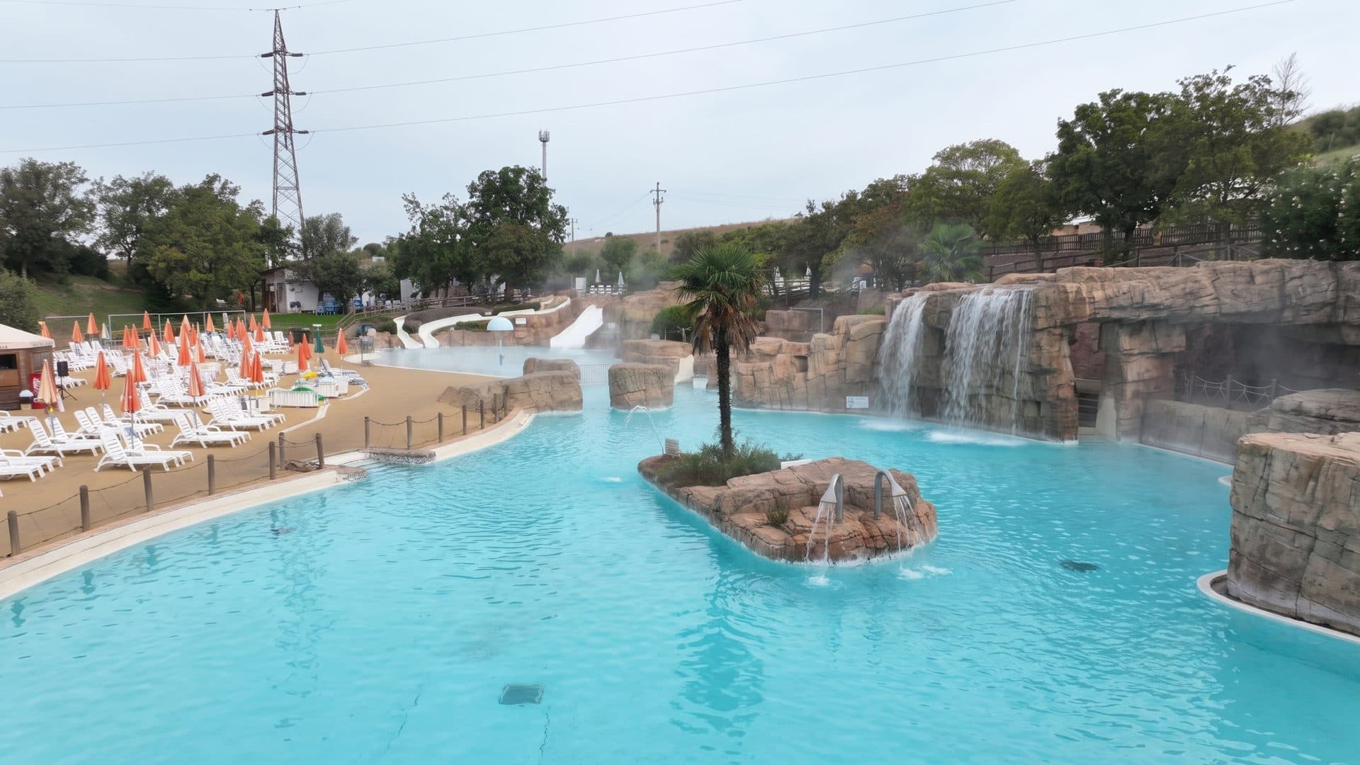 The rock-walled lagoon at Aquafelix with waterfalls spilling into turquoise water, a palm-topped island in the middle and orange loungers along the shore