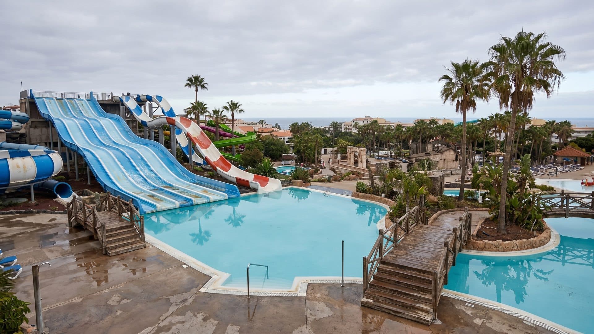 Blue and white multi-lane racing slides running down into a curving pool, with palm trees and timber footbridges