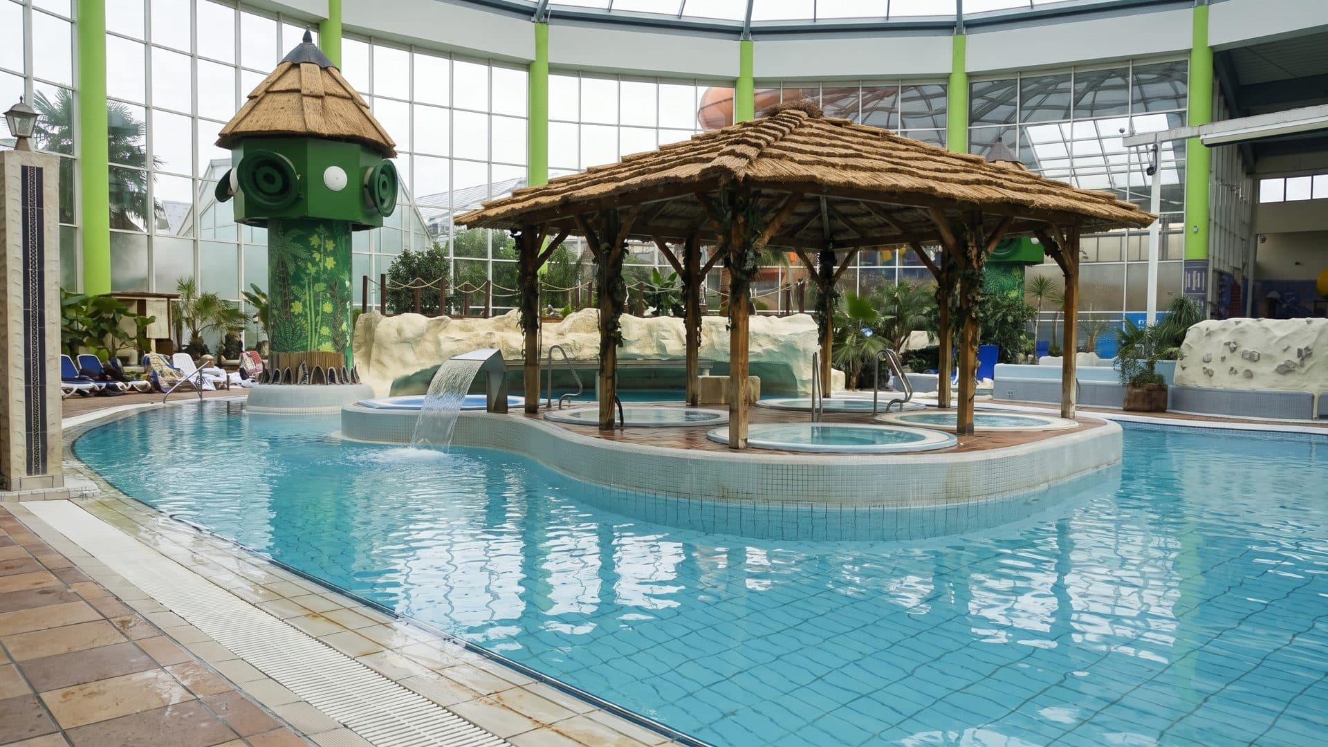 Indoor water park pool under a glazed roof, a thatched pavilion standing on the tiled shallows.