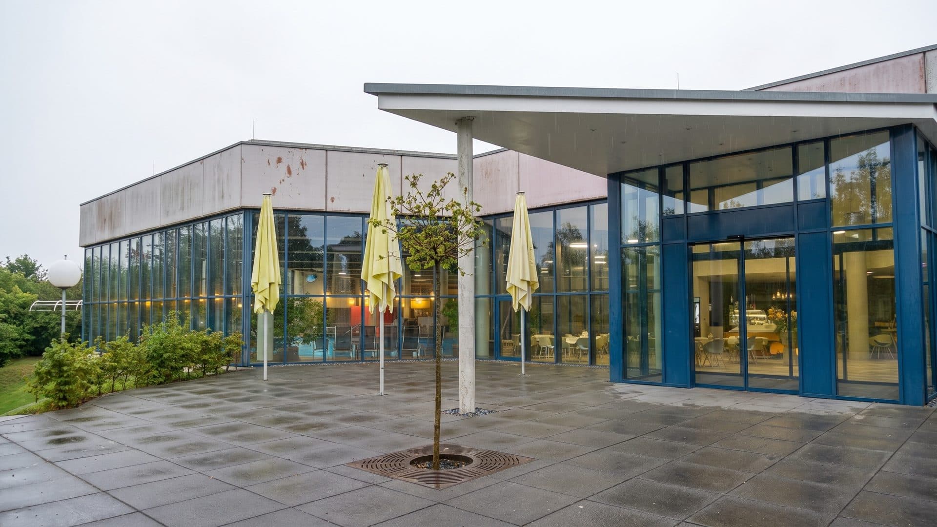 The glazed entrance pavilion and terrace of the thermal bath, with folded parasols and a young tree
