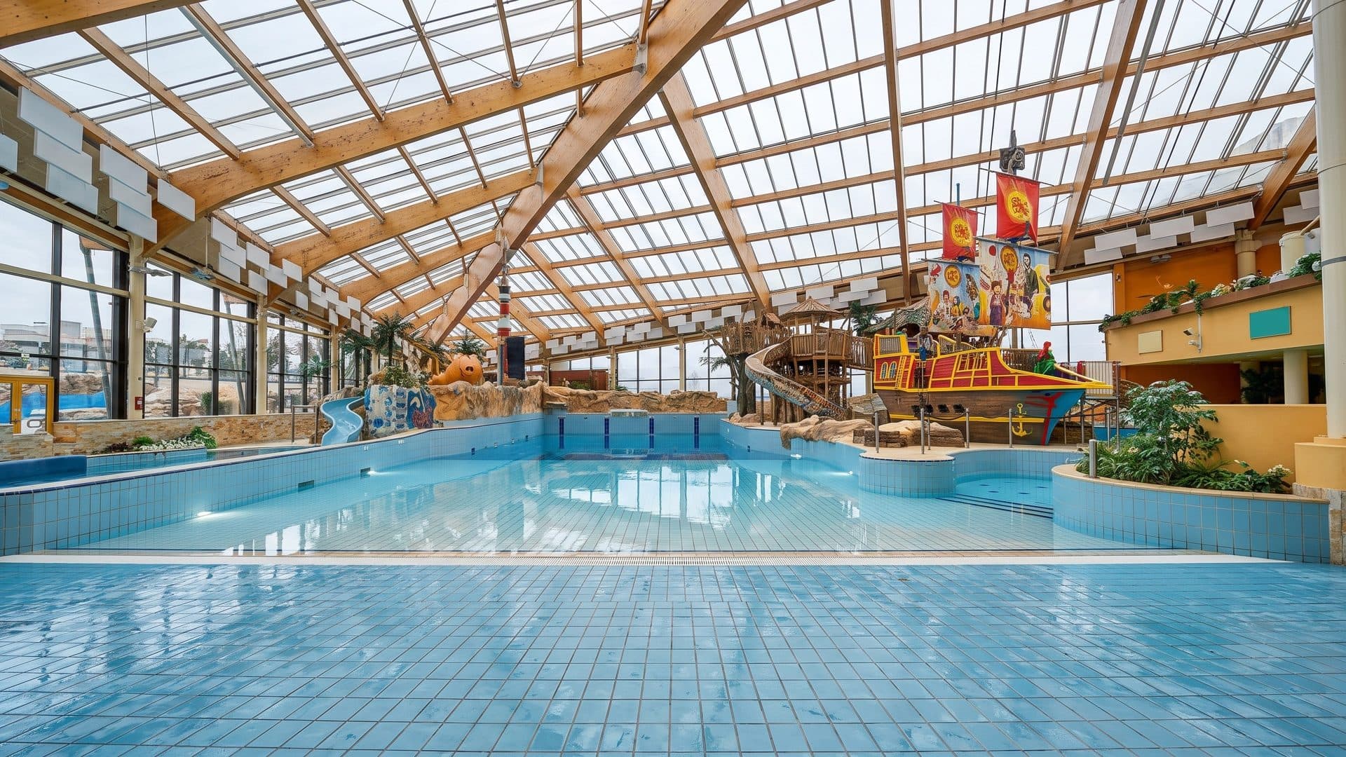 Large indoor pool hall under a timber-framed glass roof, with a pirate-ship play structure at the far end