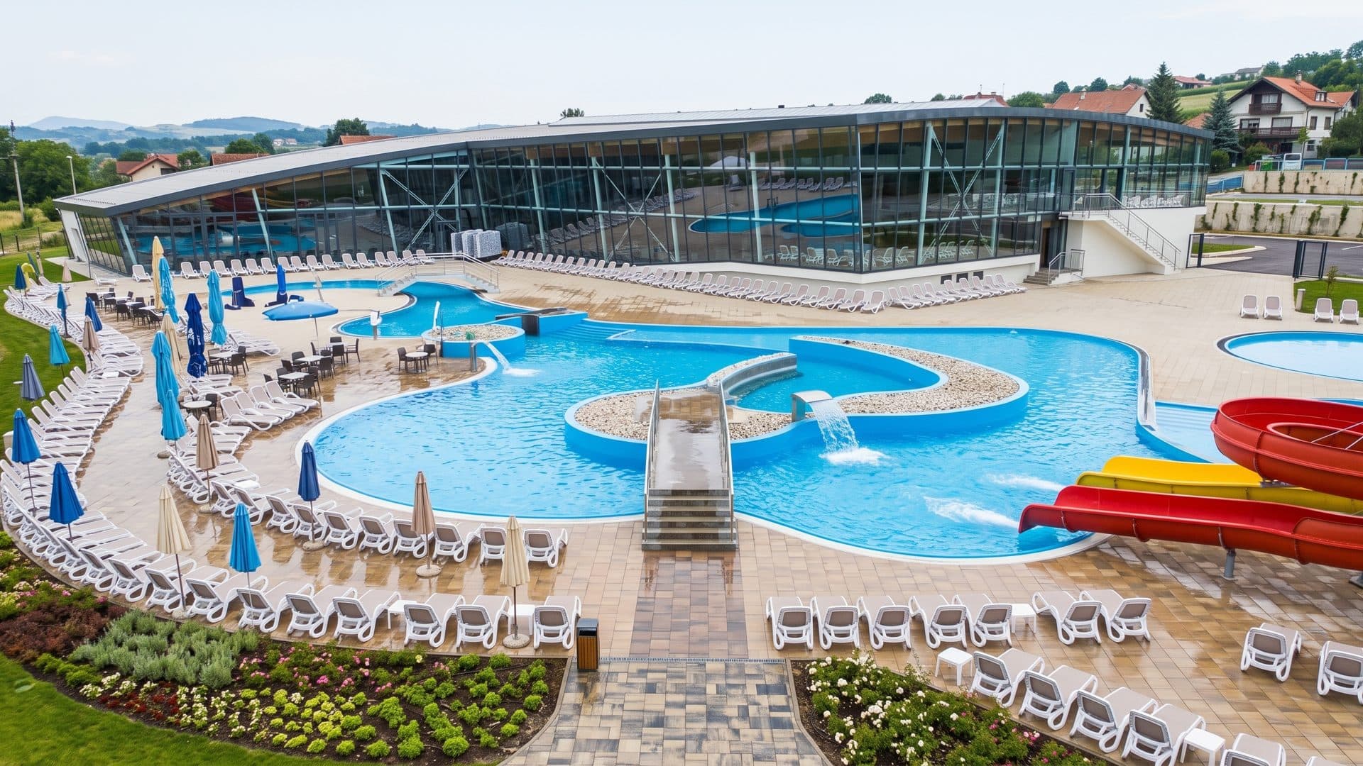 Outdoor pools with a fountain and a red slide beside the glazed hall of the water park.