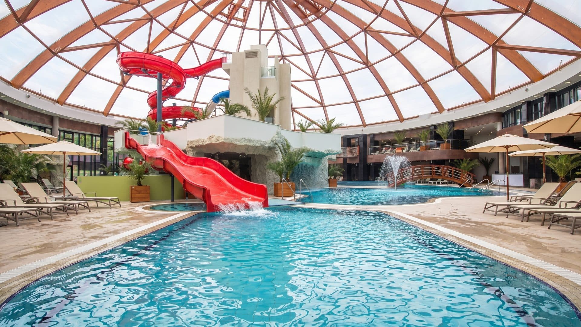 The glazed dome over Aquapark Nymphaea, a red slide curling down to the blue indoor pool with fountains and palms around it