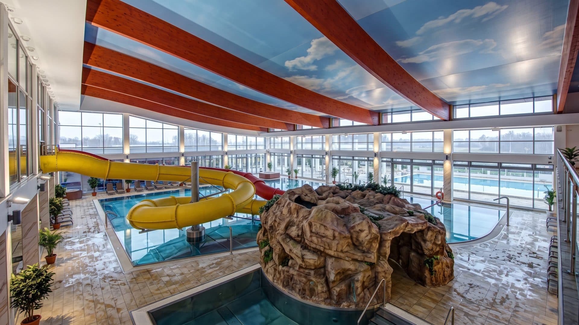 The indoor leisure pool of Aquapark Panorama Morska with a yellow slide and an artificial rock, under a painted sky ceiling