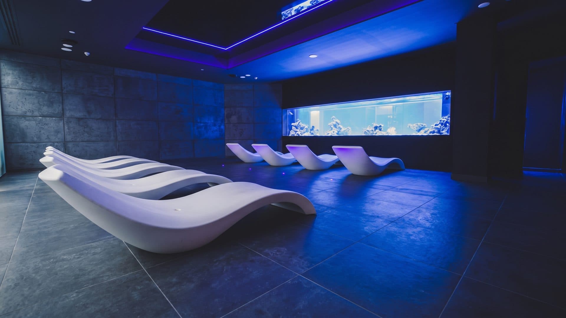 Relaxation room lit blue, moulded loungers in a row facing a long illuminated aquarium window.