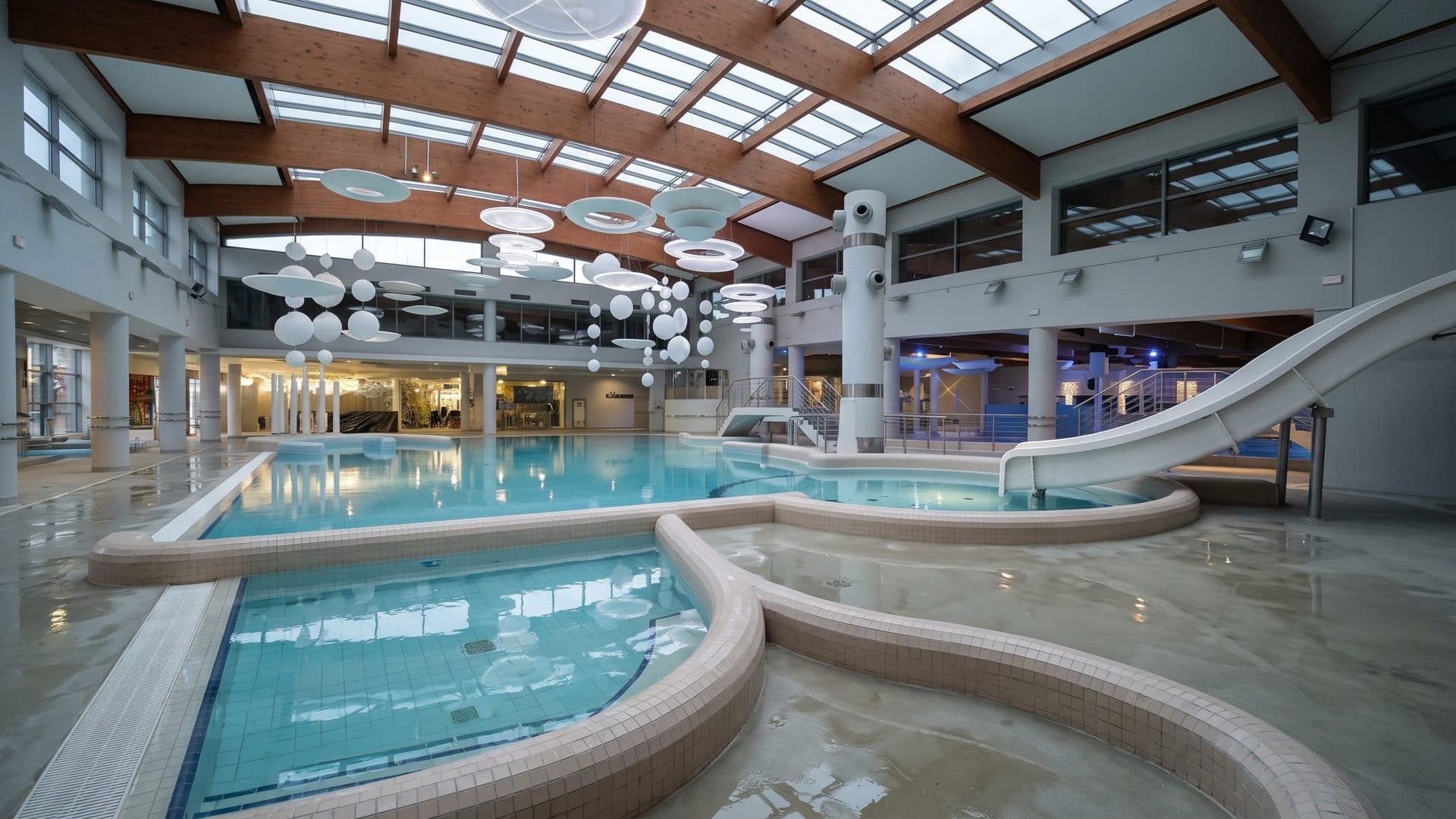 The indoor leisure pool of Aquapark Sopot with a white slide, under a timber ceiling with round lamps