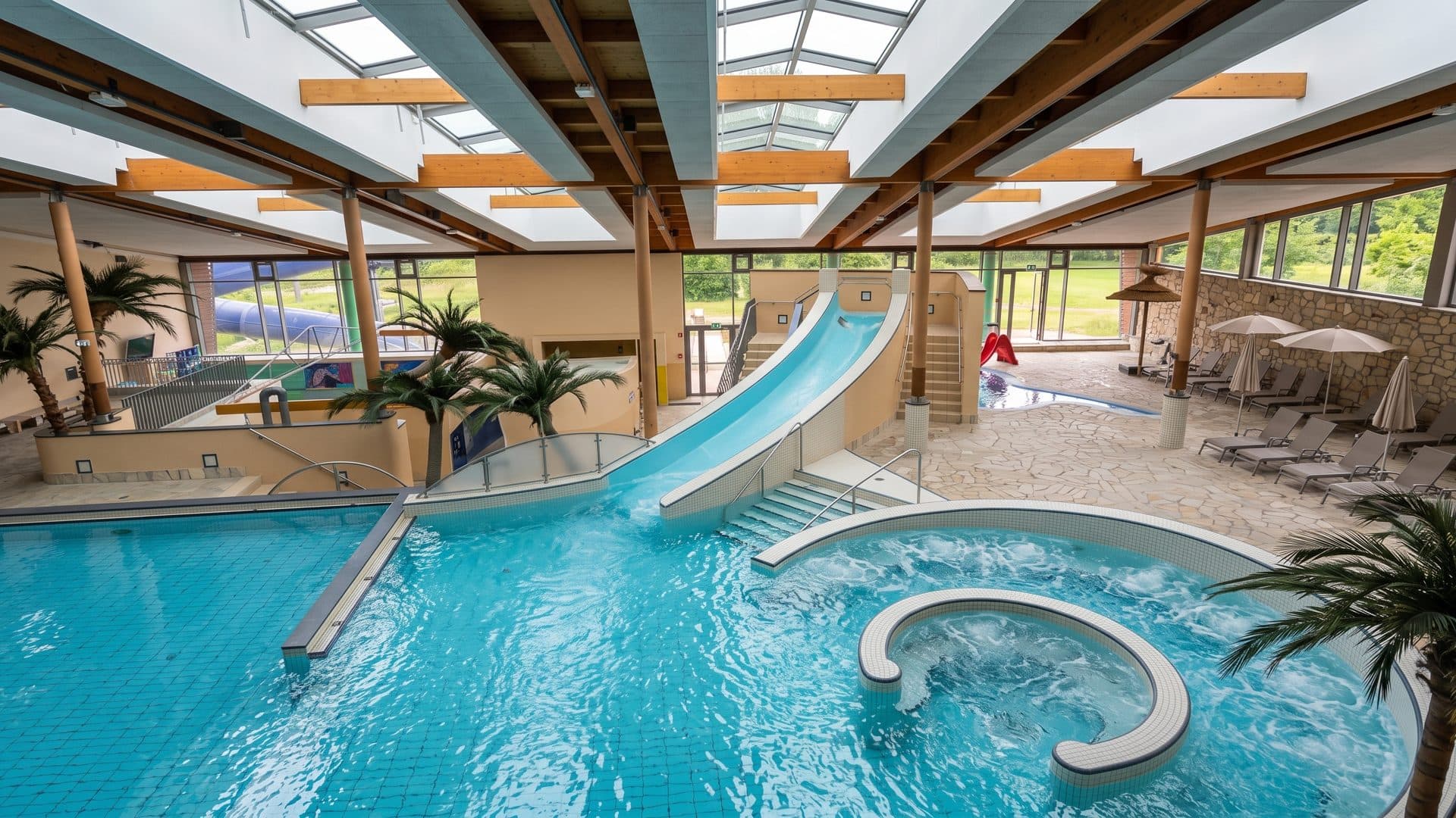 Indoor leisure pool with a blue water slide and palm planting under the glazed roof.