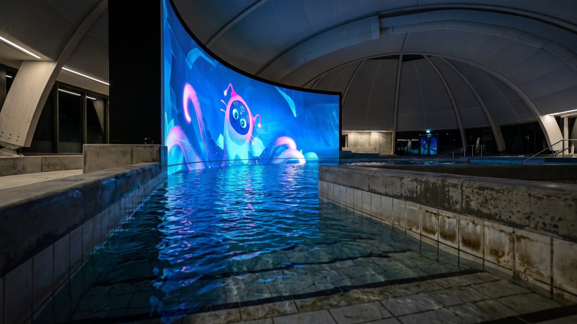 Indoor pool beneath a curved screen throwing an underwater projection across the dark hall.