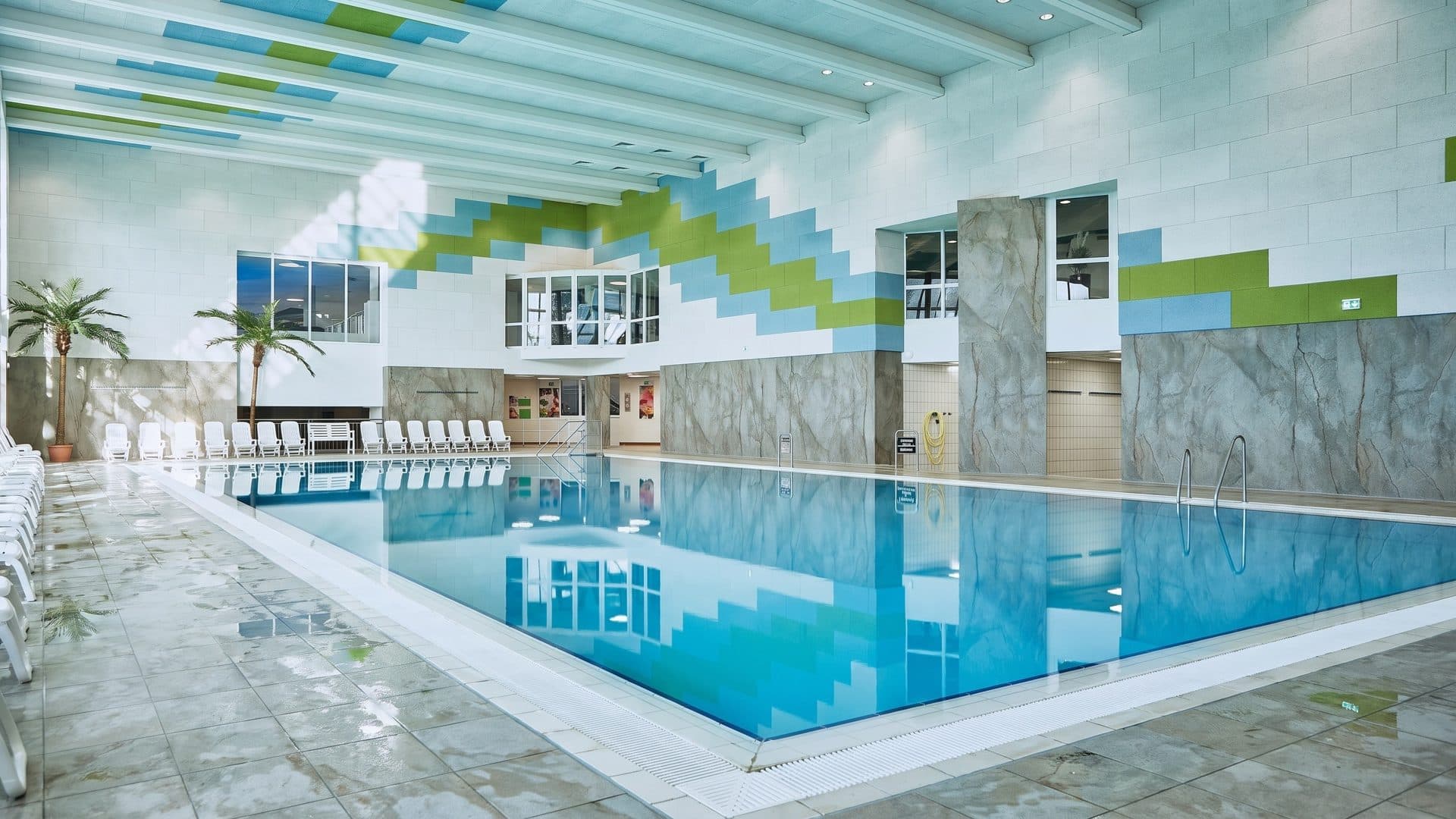 Indoor lap pool in a bright hall with a blue and green zigzag frieze and loungers along the glazed side