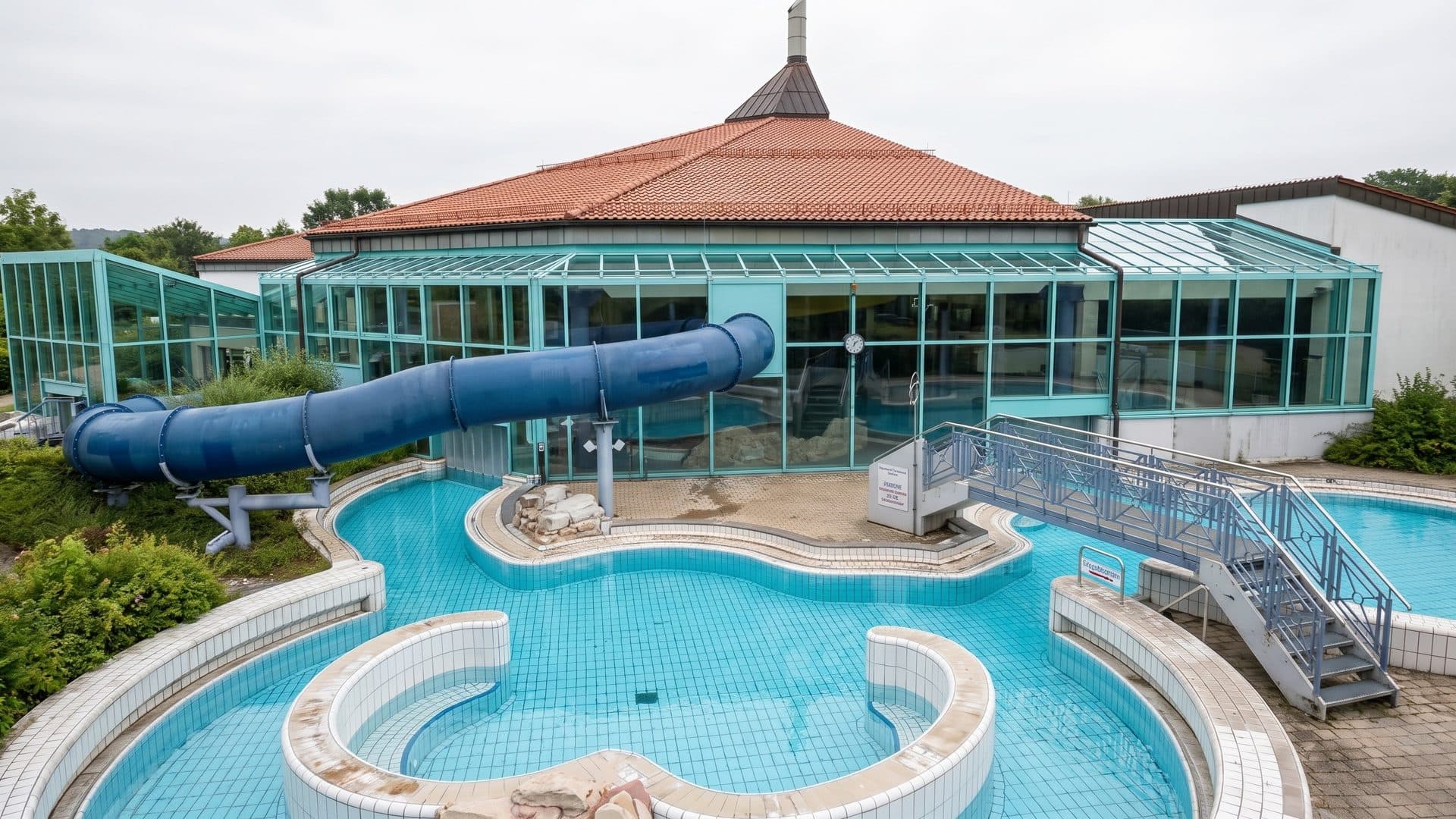 Outdoor leisure pool with round islands and a blue tube slide, in front of the glazed pavilion with a red tiled roof