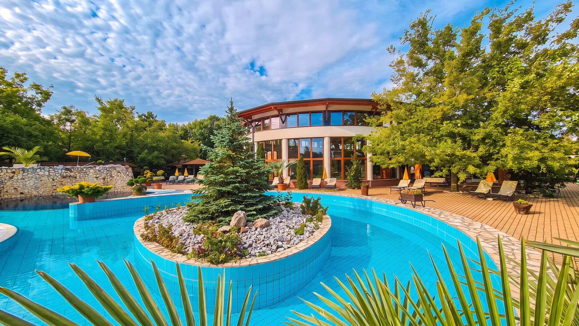 Outdoor turquoise pool curving around a planted rock island, with a wooden deck and a round glass pavilion