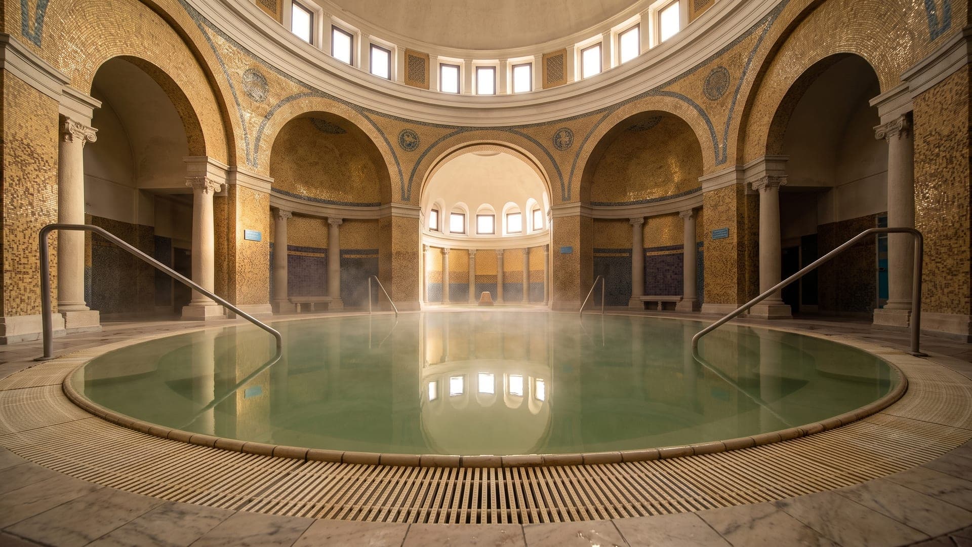The domed rotunda at the Arpad bath in Bekescsaba, its circular pool of pale green mineral water ringed by arches set with gold mosaic, steam drifting over the surface.