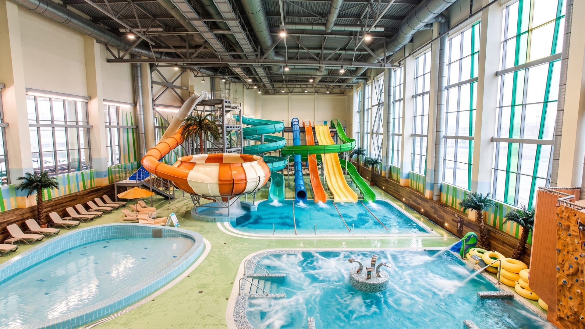 Indoor pool hall under a steel-framed glass roof, with the coloured tube slides and palm planting around the water.