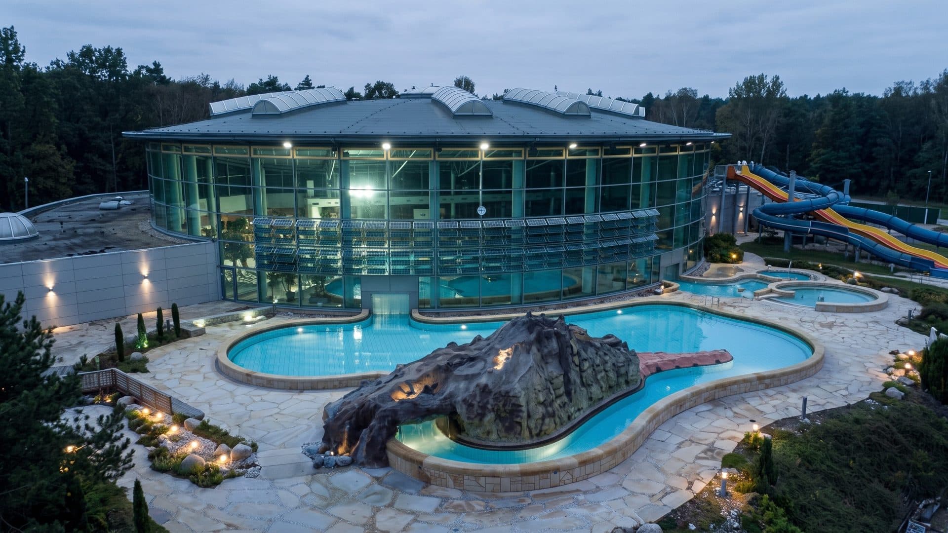Outdoor pools of the Avenida-Therme around an artificial rock at dusk, the round glass hall behind