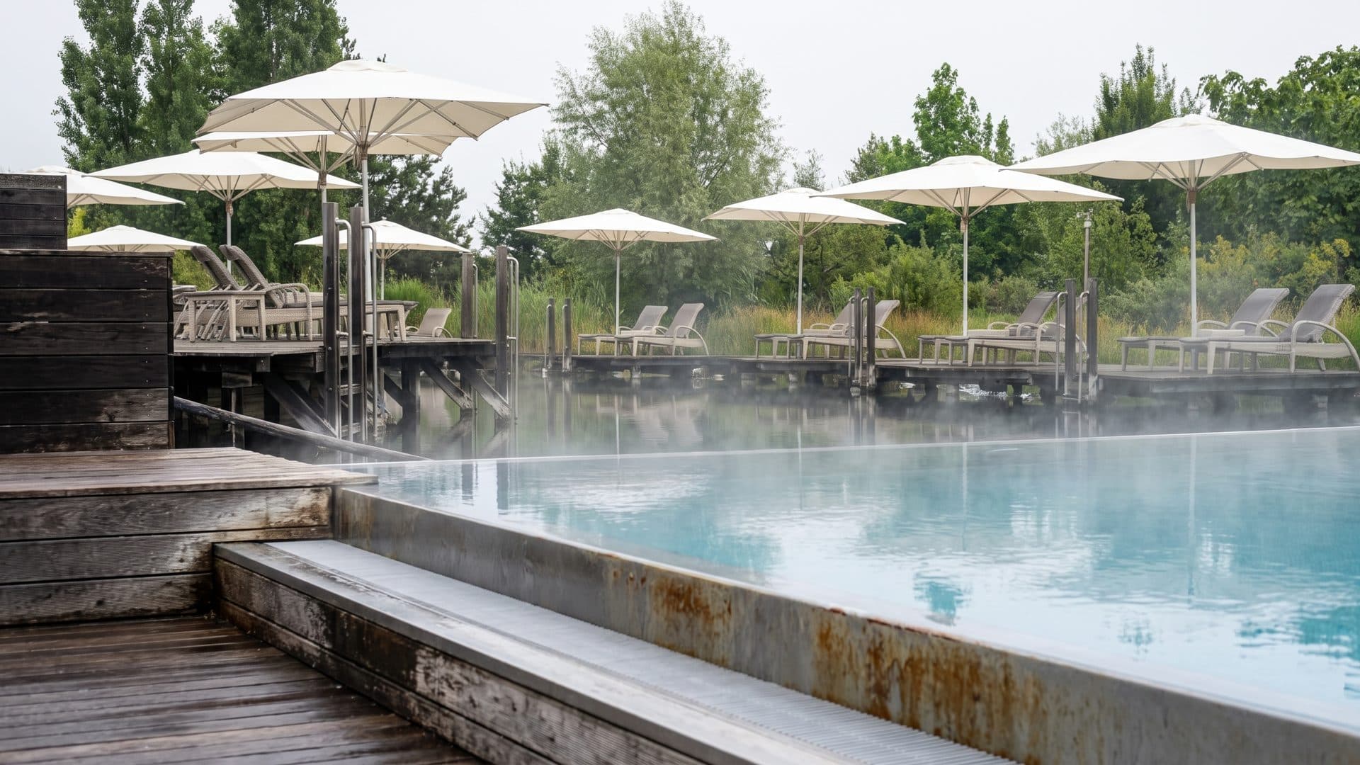 Outdoor thermal pool with steam on the water, parasols and loungers along the timber deck.