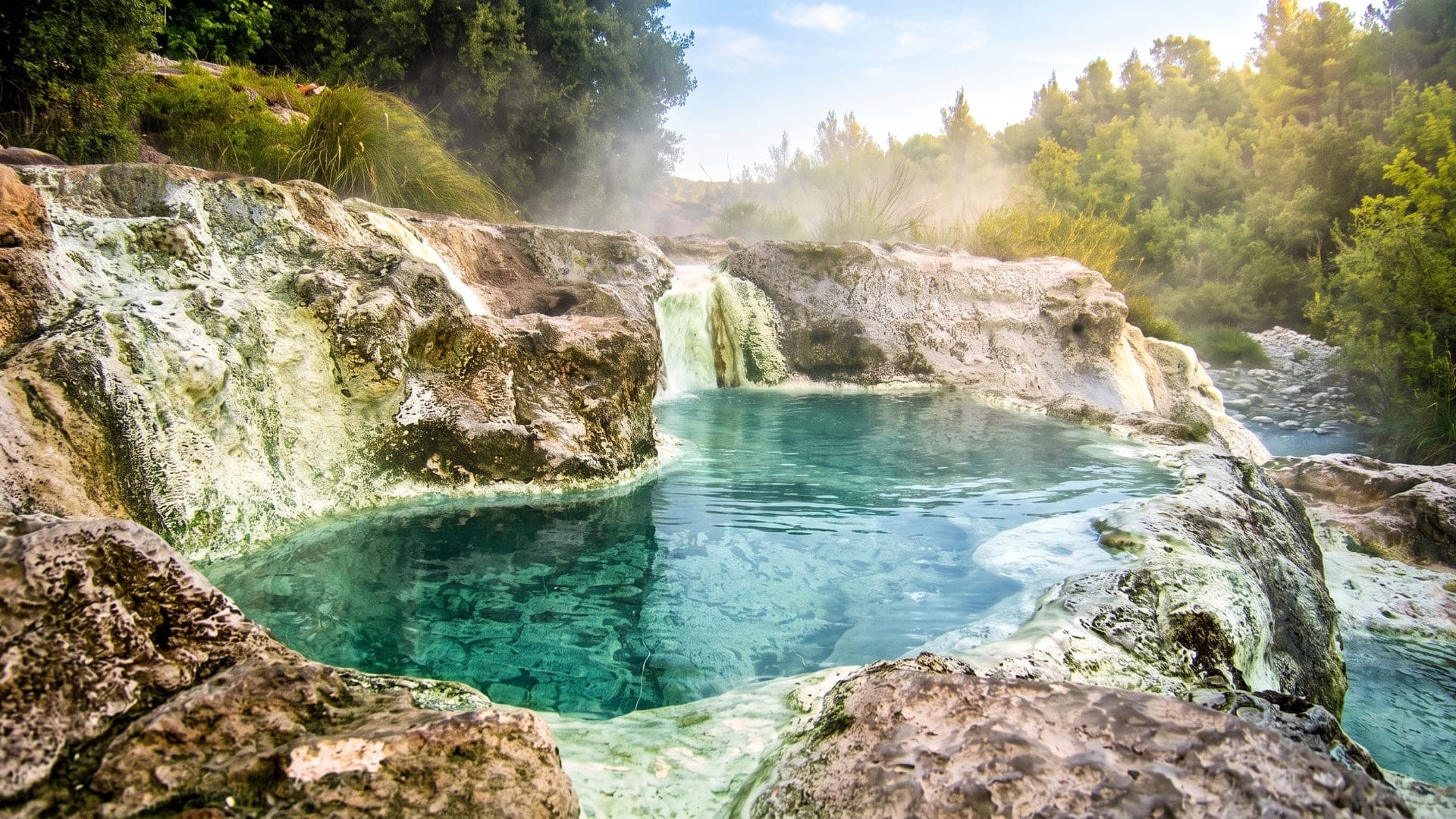 Natural travertine terraces and small waterfalls feeding a steaming turquoise pool in a wooded valley