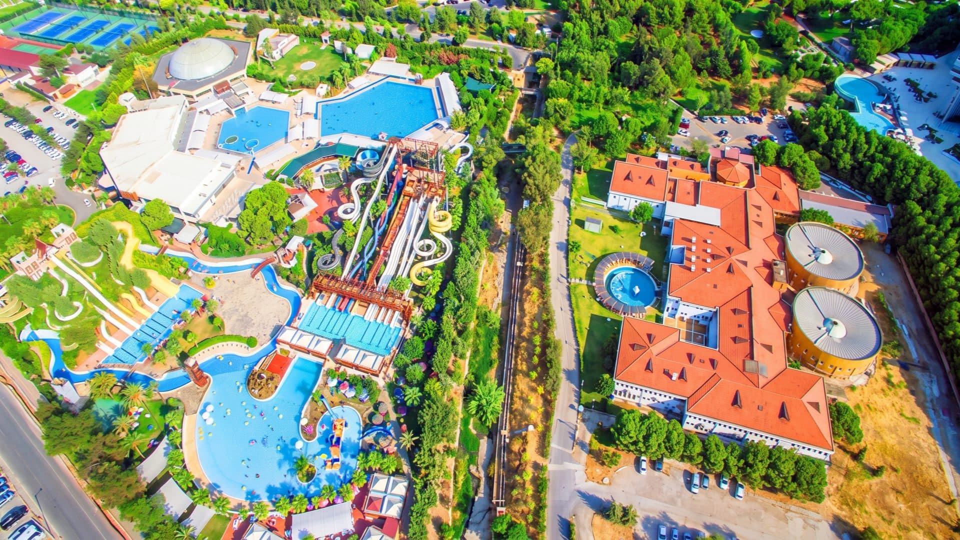 Aerial view of a large thermal aquapark, with blue leisure pools and a cluster of water slides on one side and a red-roofed spa hotel on the other, set among pine woods.