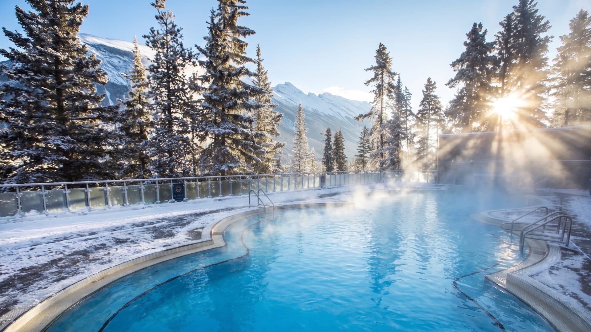 Steaming outdoor hot-spring pool in winter, its edges lined with snow and pines, a snow-covered mountain peak and low sun behind.