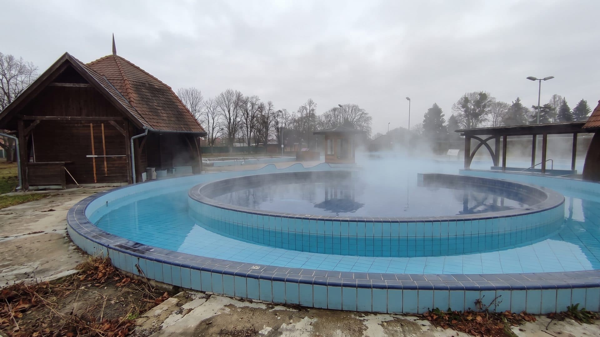 The round outdoor thermal pool at Barcs steaming in cold weather, with the timber bath building behind