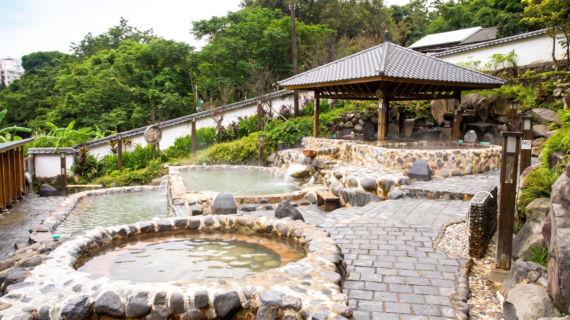 Terraced public stone hot-spring pools with pale mineral water, a tiled pavilion and dense tropical greenery on the hillside.