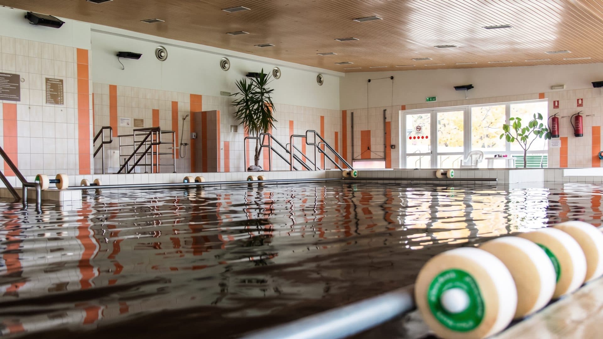 Indoor thermal pool with naturally brown mineral water, orange-striped tiled walls and aqua-aerobics floats resting on the pool edge.