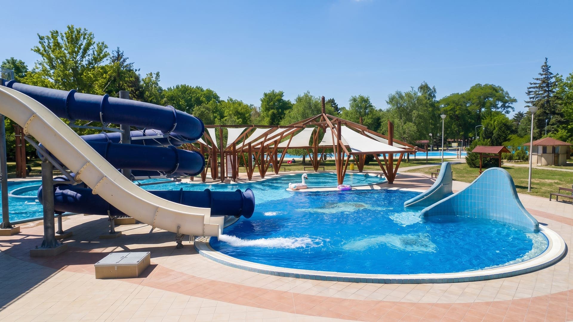 Outdoor pool with blue water slides and a timber pergola, trees beyond