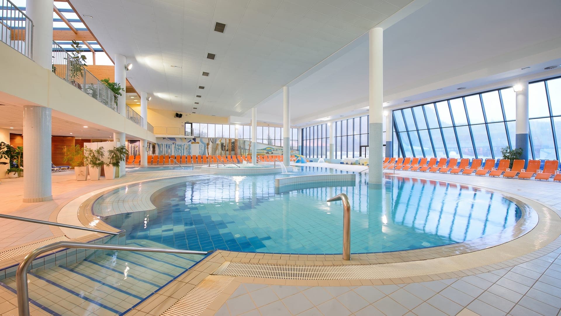 Indoor pool with a curved edge under a glazed roof, orange loungers along the glass