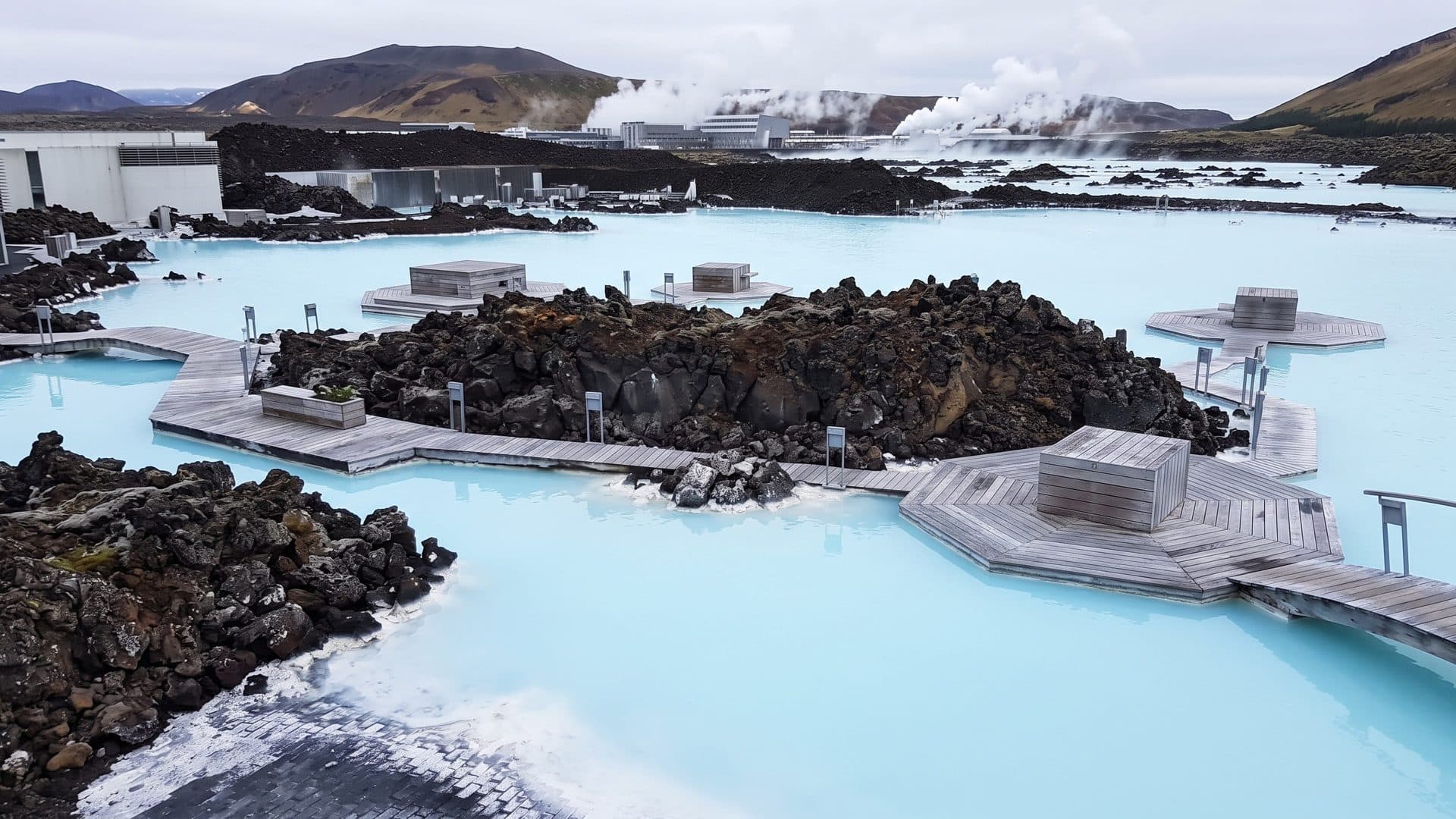 Milky blue geothermal water of the Blue Lagoon between black lava rocks and timber walkways, steam over the hills