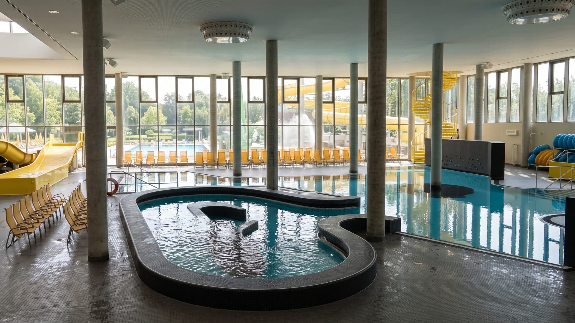 Leisure pool hall with its dark curved basin, tall windows along one side and loungers by the water.