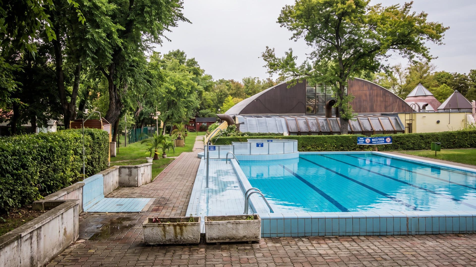 Outdoor lap pool in a leafy park, with the brown curved roof of the pool hall beyond