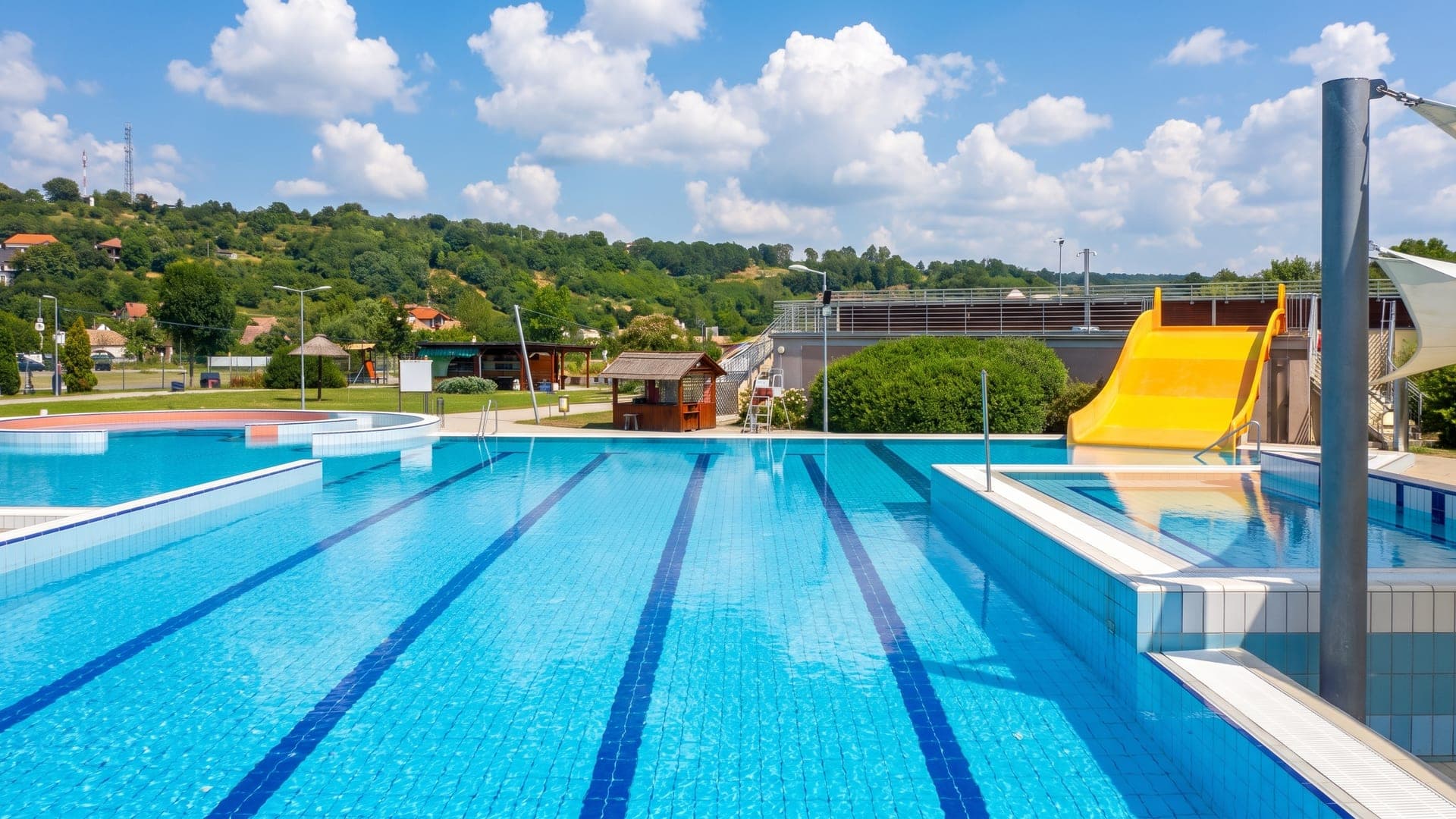 The outdoor swimming pool at the thermal bath in Bonyhad, with a yellow slide and the wooded hills of Tolna behind.