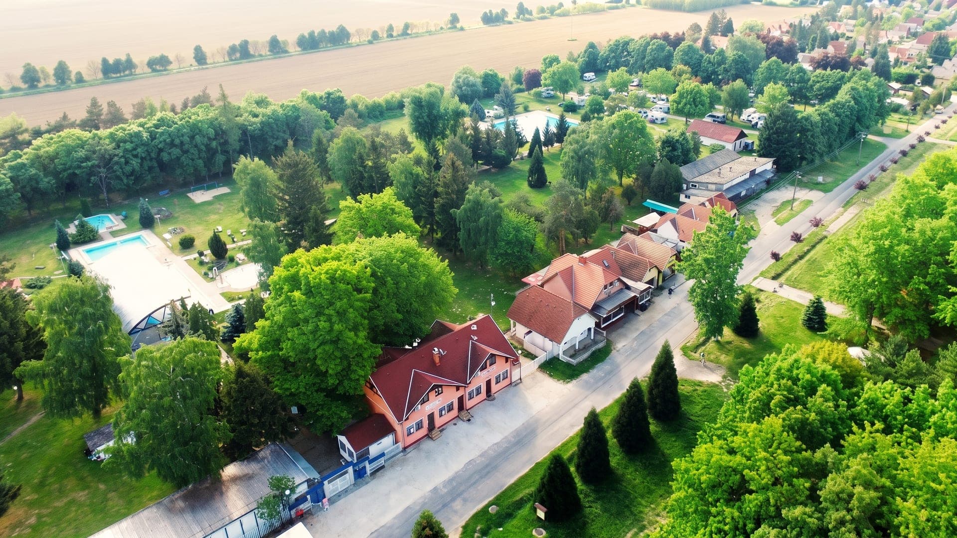 Borgata Termalfurdo seen from the air, its open-air pools among trees at the edge of the village, with fields beyond.