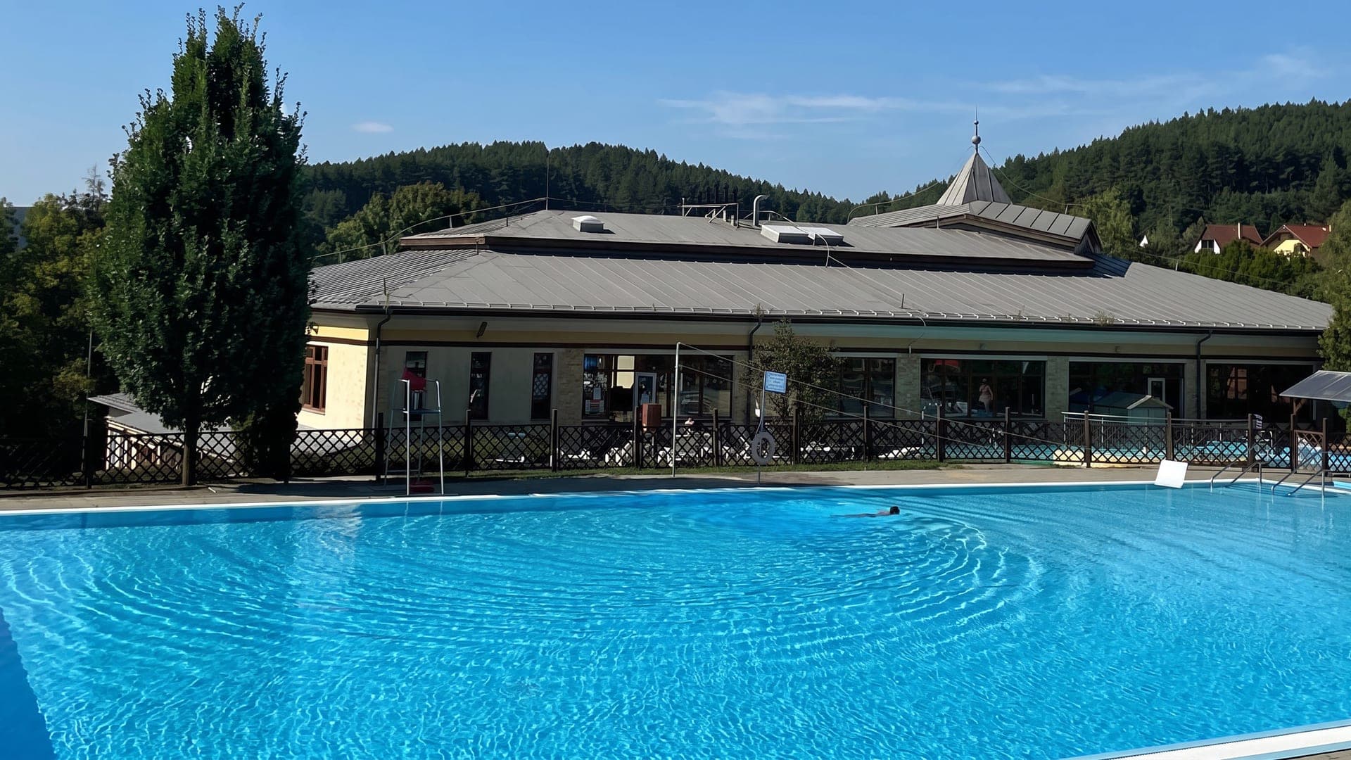 The outdoor pool at the thermal bath in Bukkszek, with the long bath house behind it and the wooded Bukk hills rising beyond.